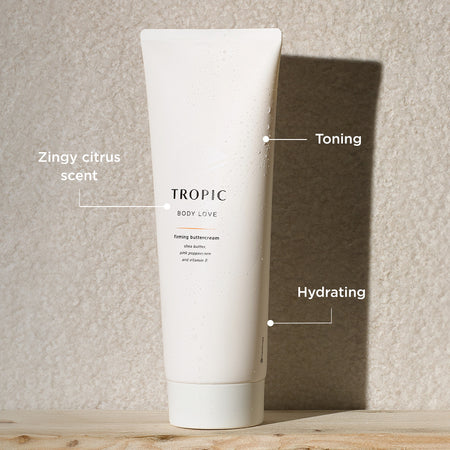 The image displays a tube of Tropic Body Love Firming Buttercream standing against a textured wall with sunlight casting a soft shadow. Key features of the product are labeled:
Zingy citrus scent, providing a refreshing, uplifting aroma.
Toning, aimed at improving skin firmness and elasticity.
Hydrating, formulated with nourishing ingredients like shea butter, pink peppercorn, and vitamin E to deeply moisturize the skin.